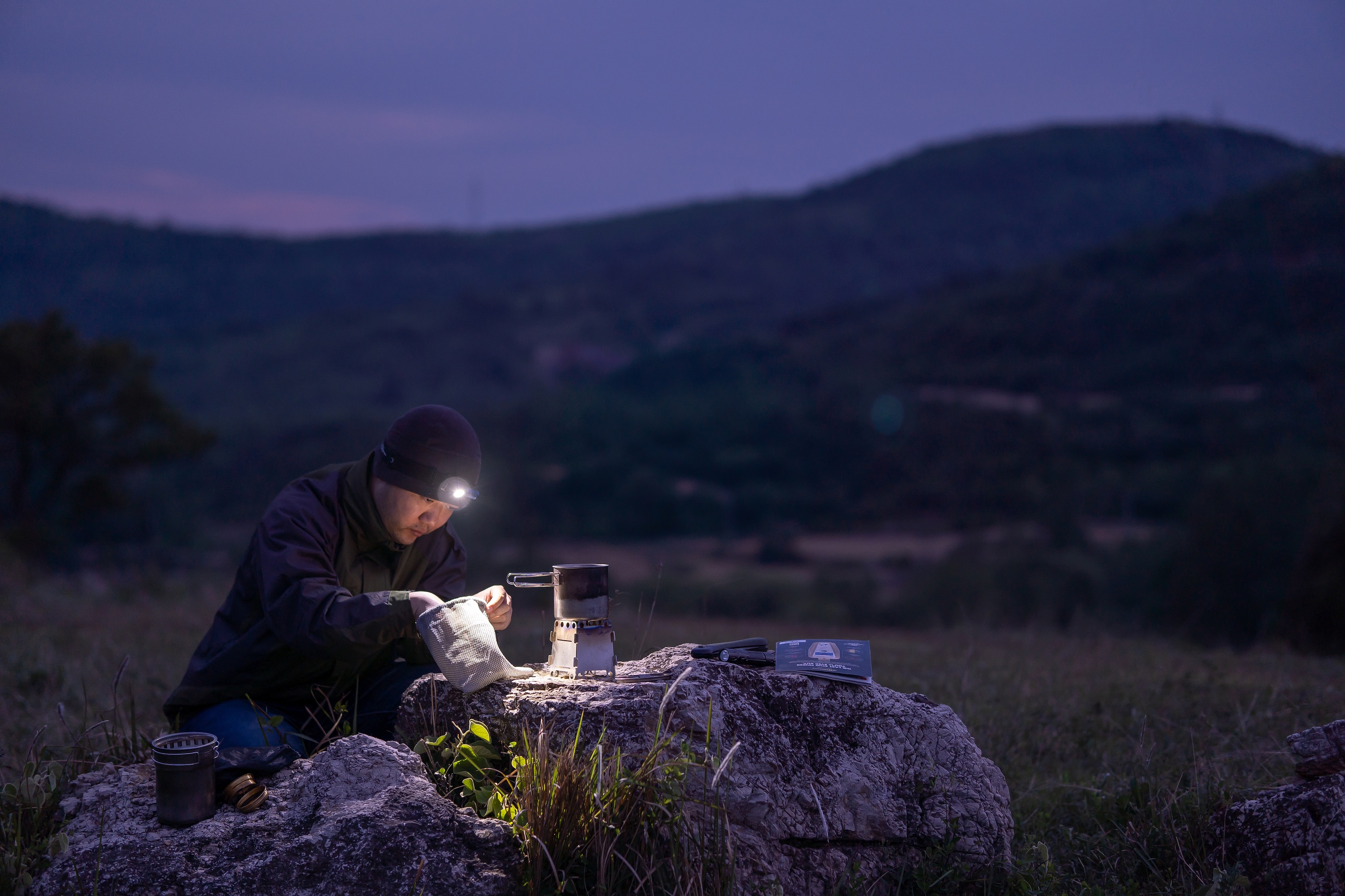 Mann mit Stirnlampe kocht nachts draußen auf tragbarem Campingkocher auf Felsen. Ideal für Outdoor, Camping, Wandern und Rucksacktouren bei Dunkelheit.