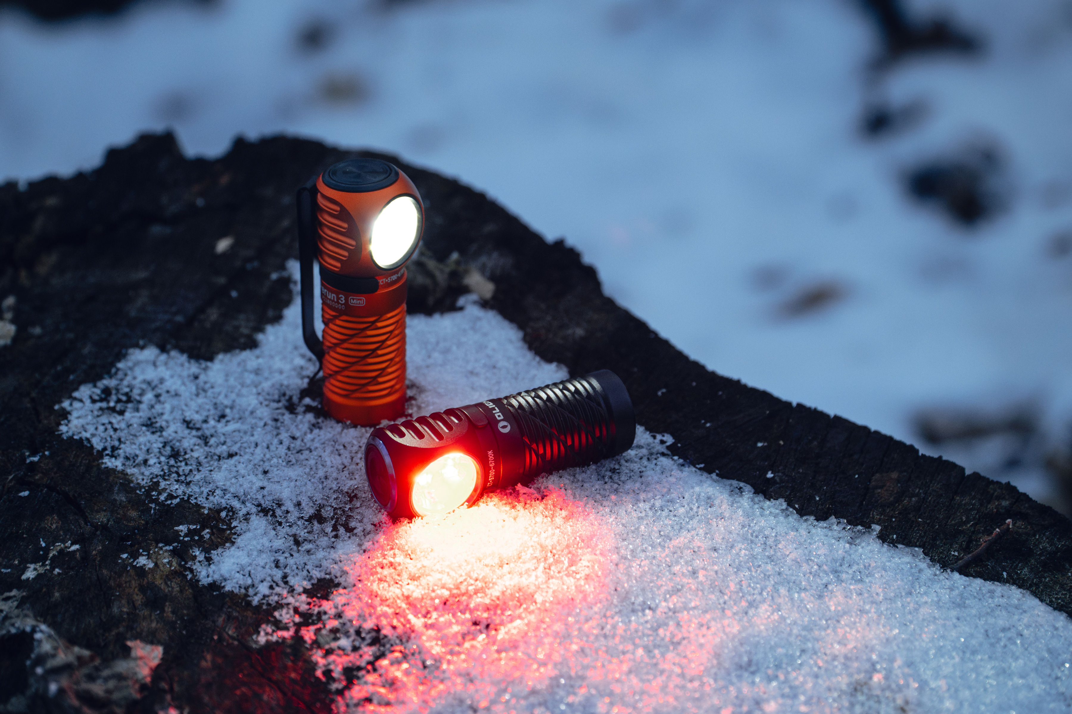 Dos linternas encendidas sobre nieve y madera oscura. Una naranja vertical emite luz blanca; la negra horizontal, luz roja.