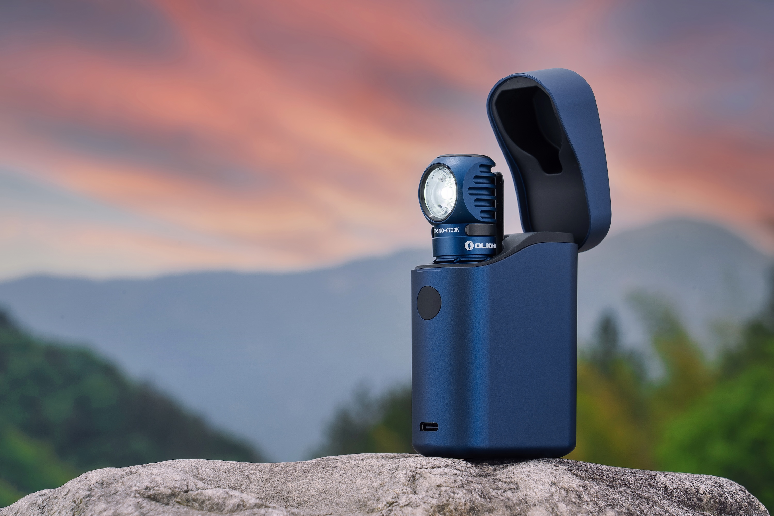 Blue Olight portable power bank with detachable LED flashlight on a rugged rock, overlooking a scenic mountain range and vibrant sunset sky. Ideal for outdoor adventures and camping.