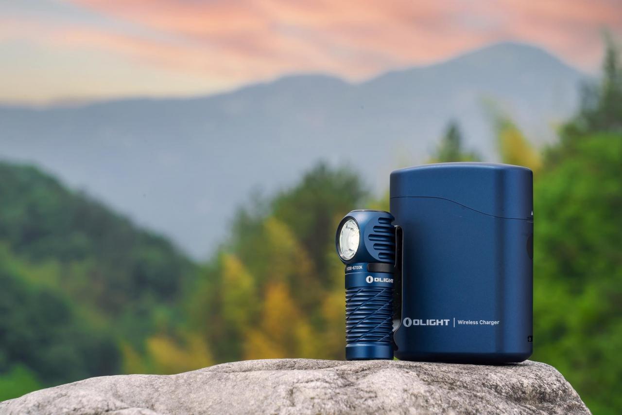 Olight blue compact flashlight and wireless charging case on a rock with a scenic mountain sunset background