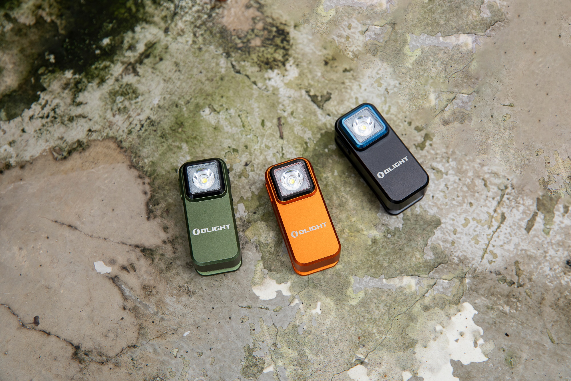 Three Olight compact rectangular flashlights in green, orange, and black, displayed on a textured, earthy surface.