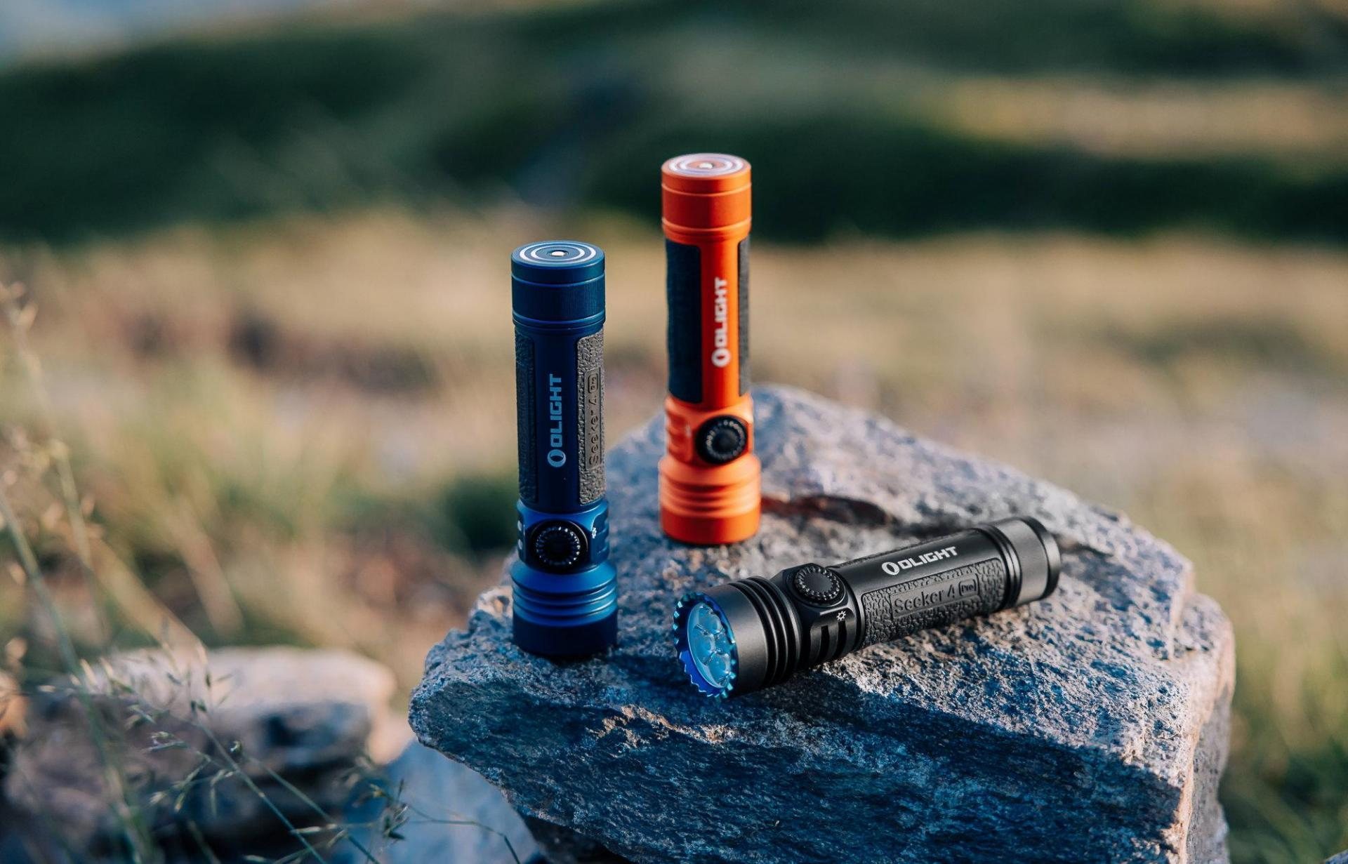 Three Olight Seeker 4 Pro flashlights (blue, orange, black) on a rock outdoors. Durable LED lights for hiking, camping, and tactical use.