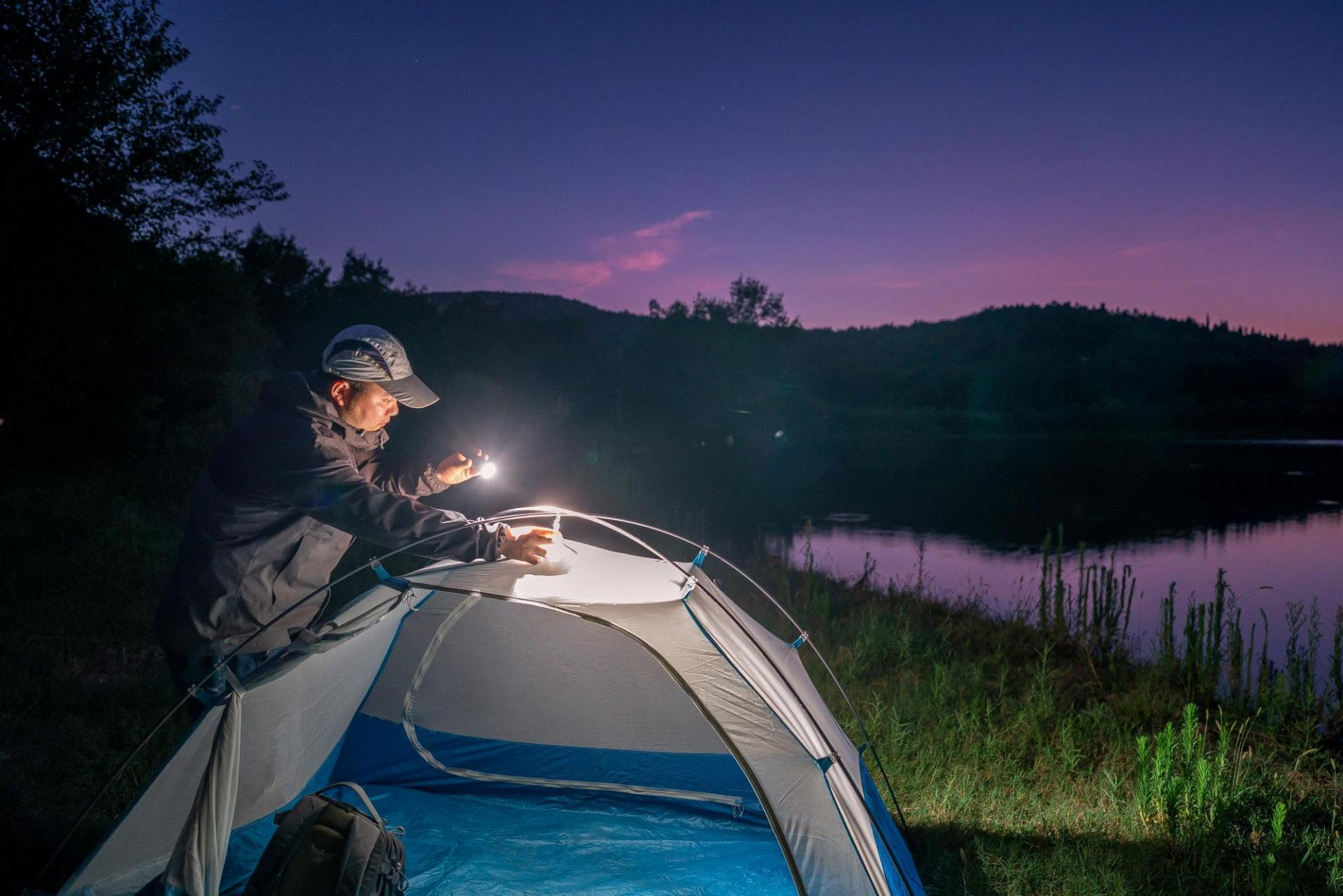 Man lighting up a camping tent with a flashlight at dusk by a lake, an outdoor adventure scene.
