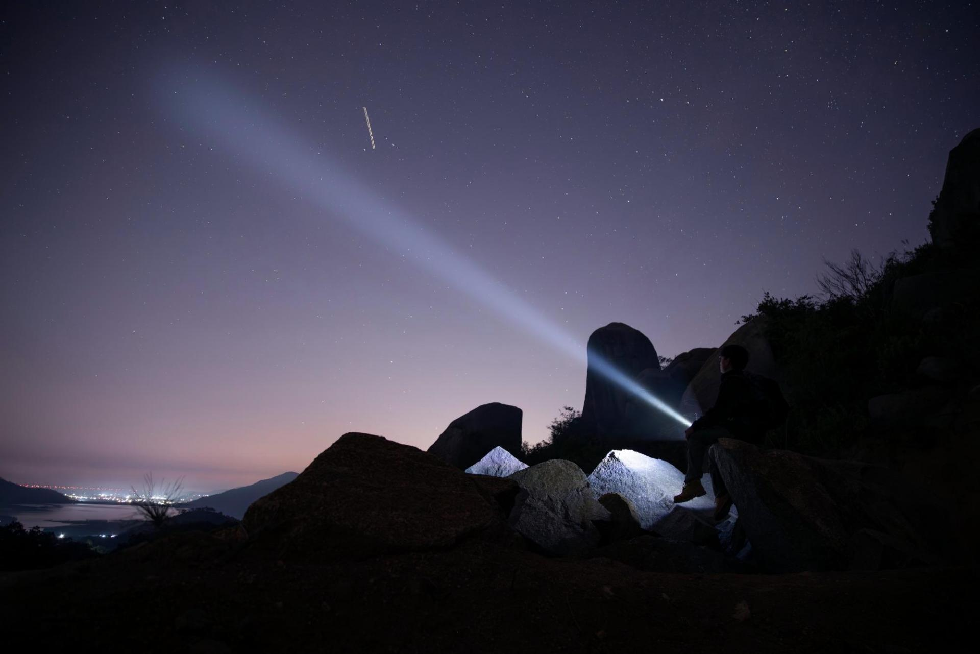 Person sitting on rocks, shining a bright flashlight beam into a star-filled night sky above distant city lights and mountains. Night exploration and stargazing scene.