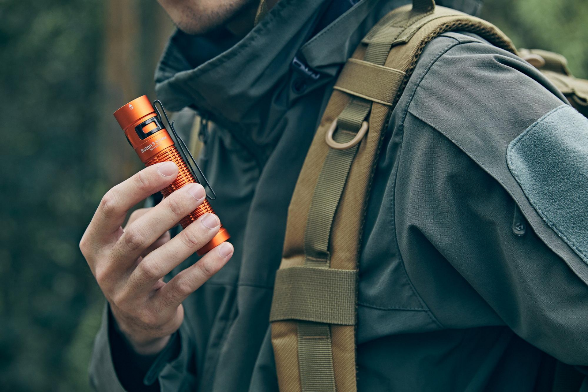 Olight Baton 3 Pro Max orange flashlight held in hand, showing tactical outdoor gear and a backpack. Ideal for hiking, camping, and EDC.