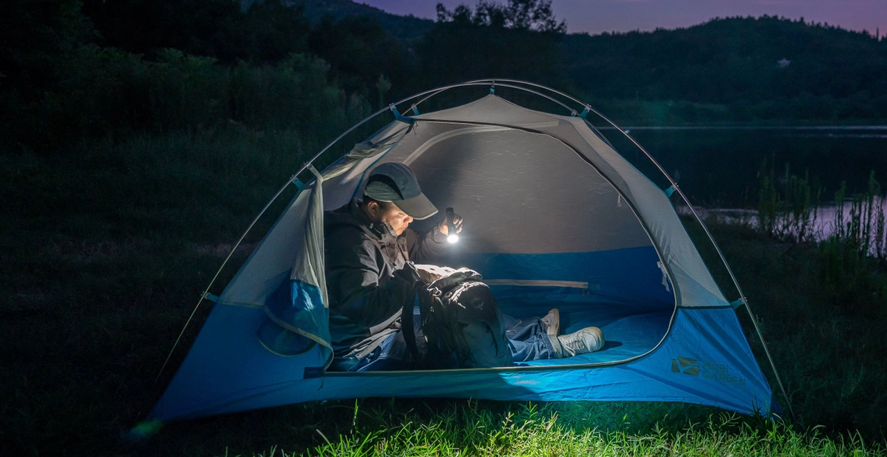 A man using a portable flashlight to search his backpack inside a blue camping tent at night by a lake.