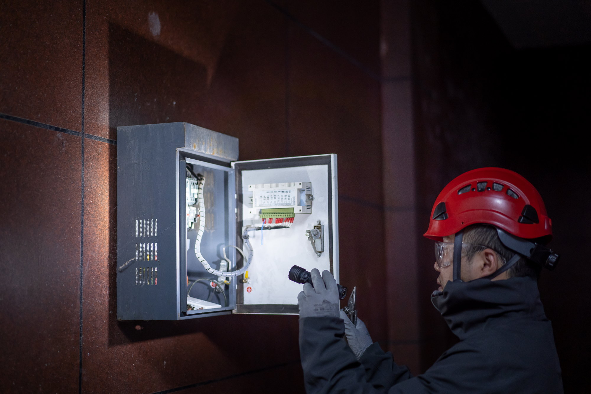 Technician in hard hat inspecting an electrical control panel with a flashlight