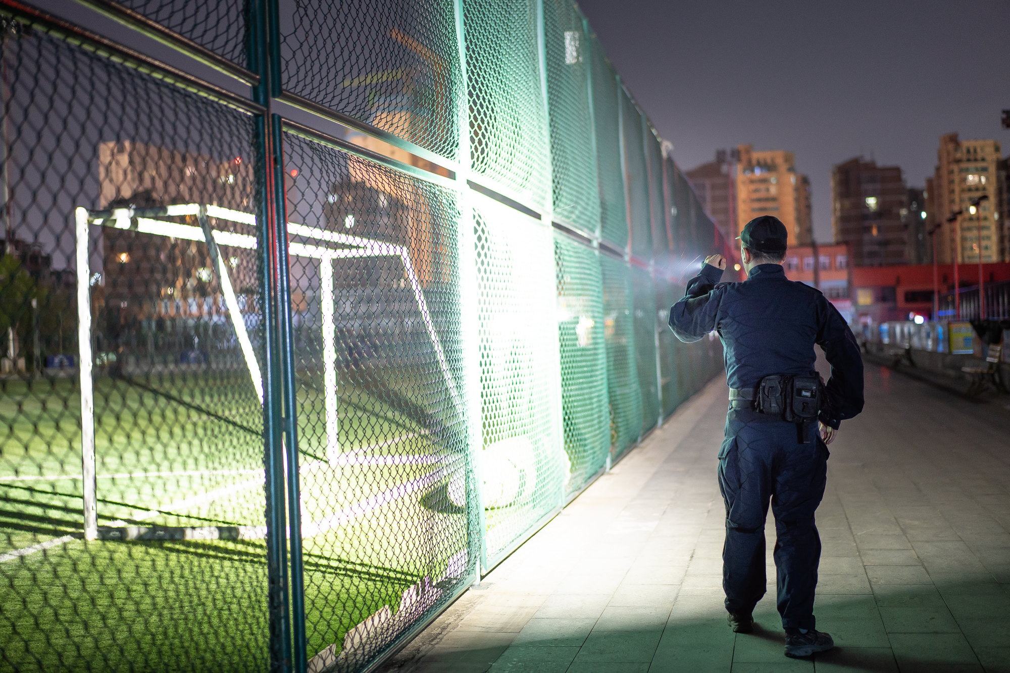 Security guard shining flashlight on a fenced soccer field at night