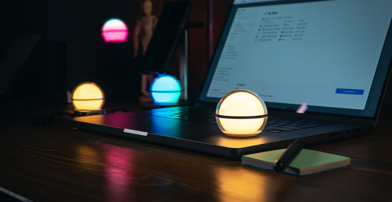 Lámparas LED esféricas de colores iluminan un escritorio con portátil, notas adhesivas y bolígrafo, creando un ambiente de trabajo moderno y acogedor.