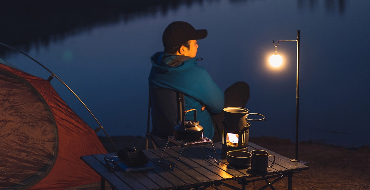 Solo man camping by a calm lake at night, sitting next to a tent with camping gear, a portable stove, kettle, and a warm hanging lantern on a folding table.