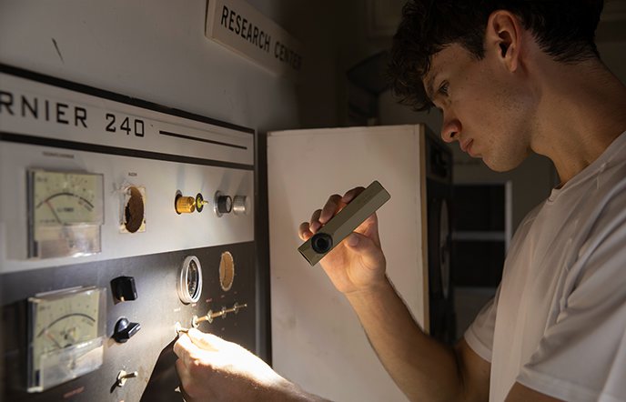 A researcher using a flashlight to inspect a vintage industrial control panel labeled RNIER 240 in a research center, highlighting analog dials and switches.