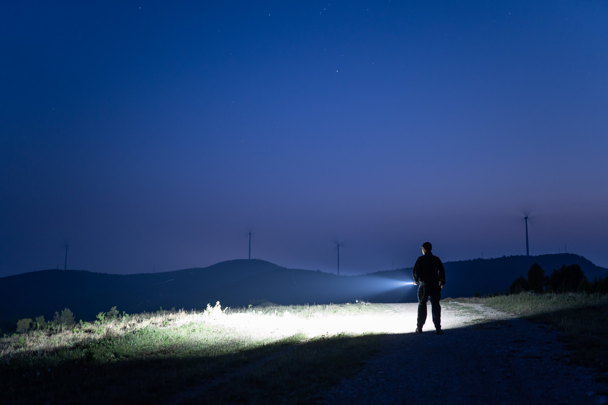 夜の山道で強力なLED懐中電灯を使い、前方を明るく照らす男性のシルエット。背景には美しい星空と風力発電の風車が広がる、キャンプや夜間活動、防災対策に最適な高輝度フラッシュライトの利用シーン。