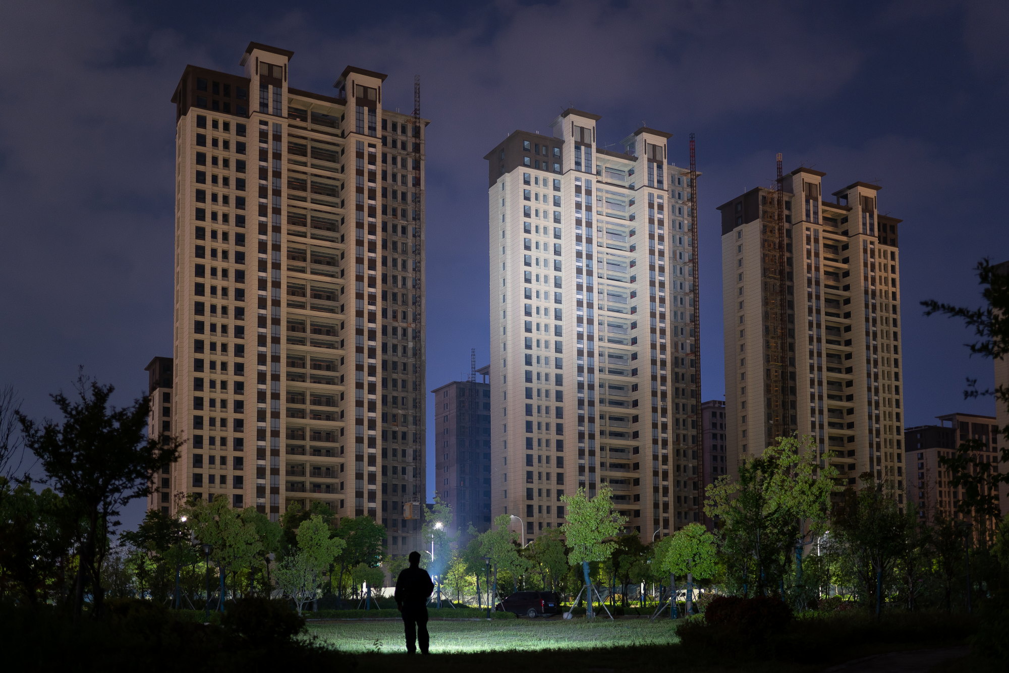 夜間にライトアップされた高層マンション群と、手前の公園に立つ人物のシルエット。都市の静かな夜景と近代的な住宅開発の風景。