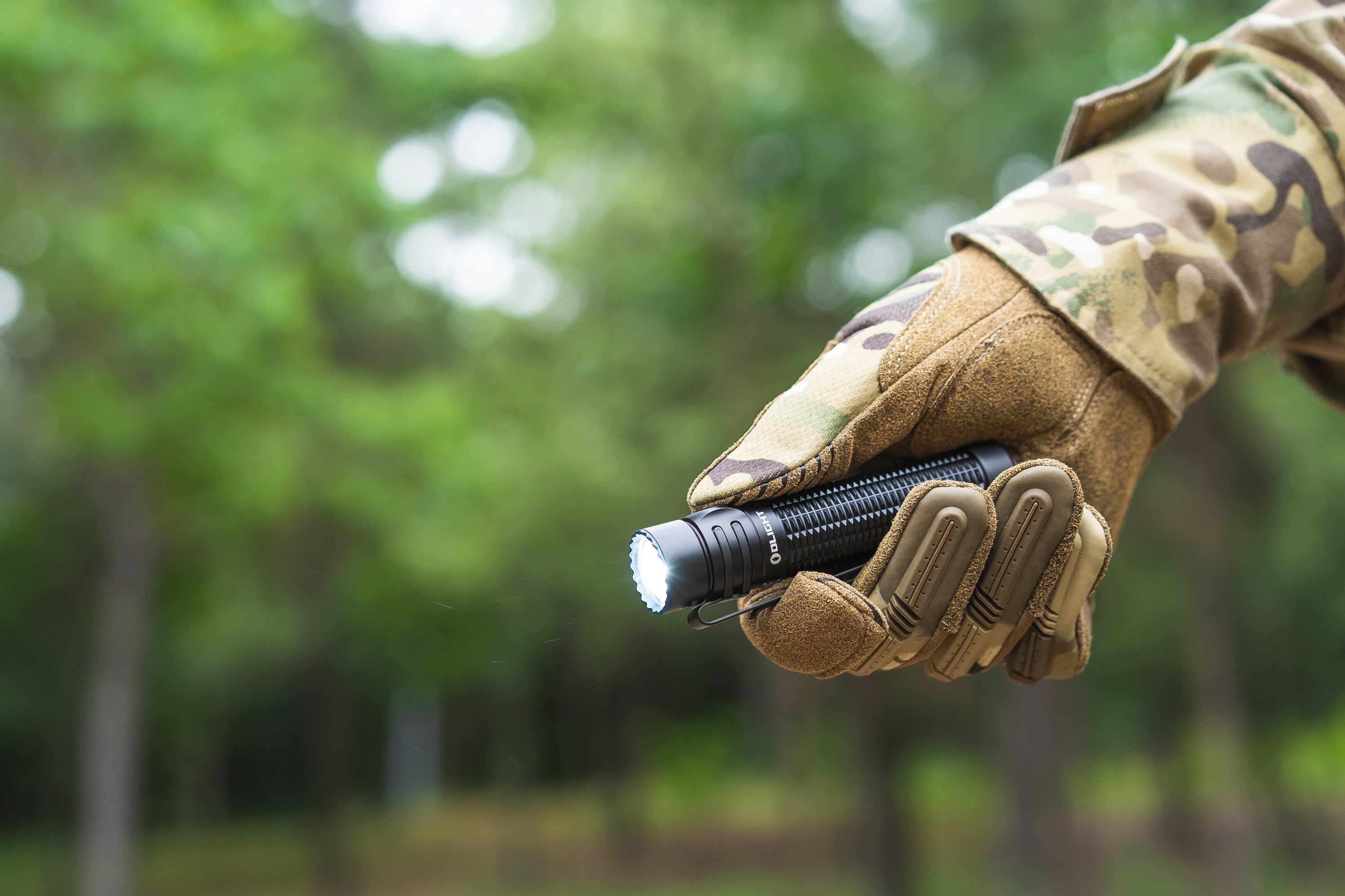 迷彩グローブをはめた手が、森を背景に明るく点灯した懐中電灯を持っている様子