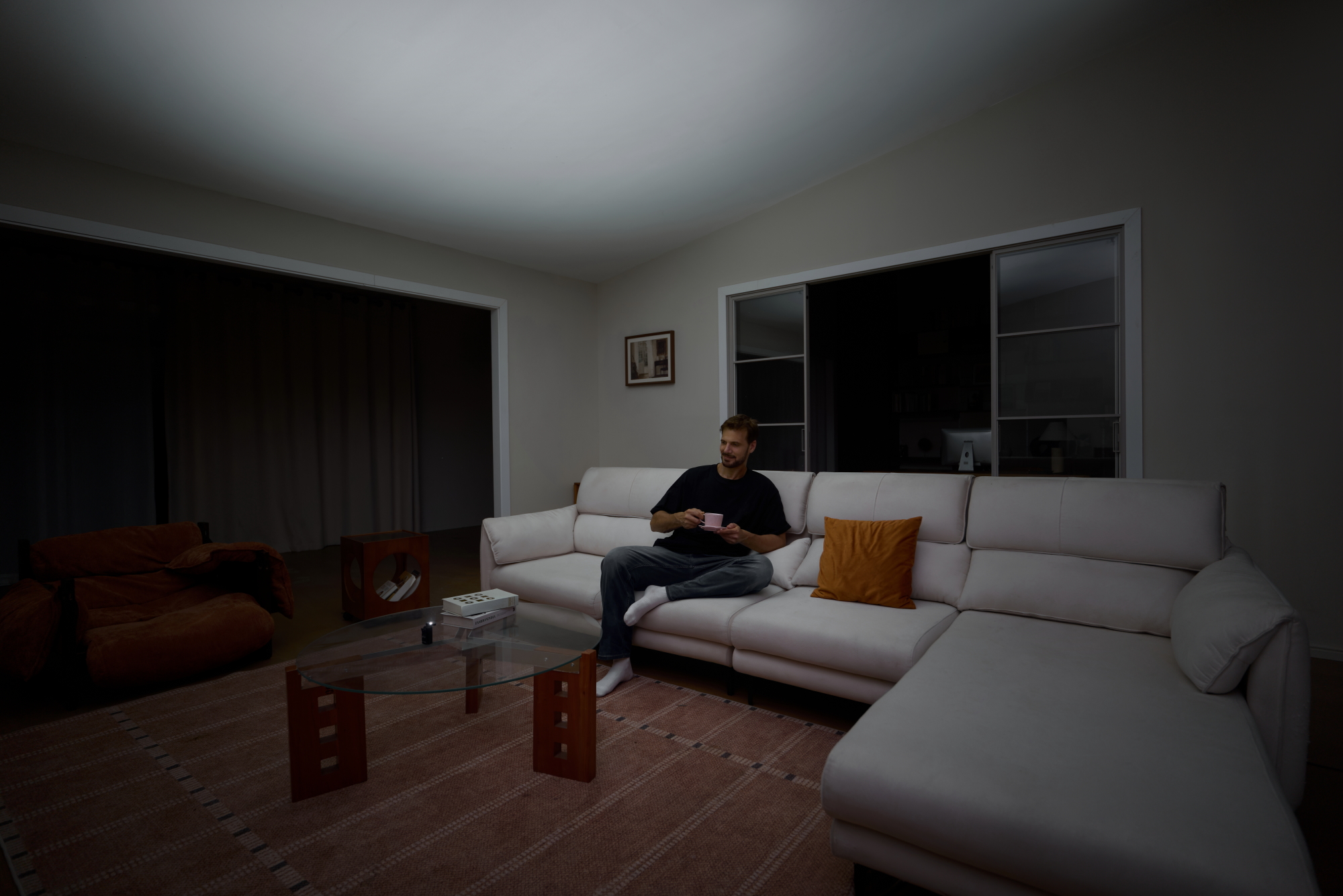 Man holding a coffee cup while relaxing on a large white leather sectional sofa in a modern, dimly lit living room at night.